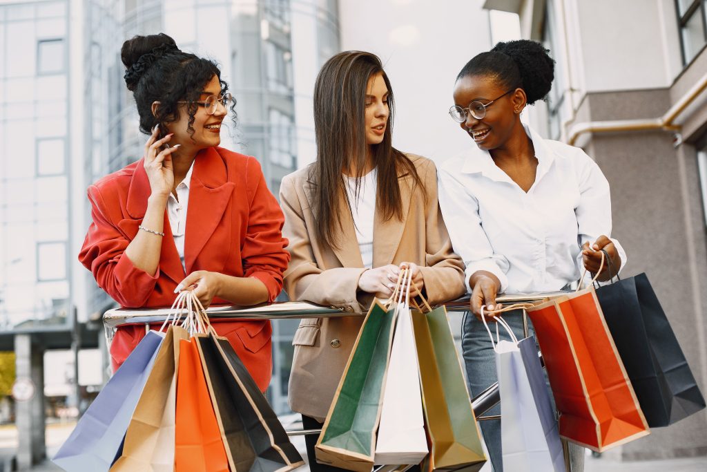 Three women with shopping bags chatting happily outdoors, showcasing diversity and fashion.